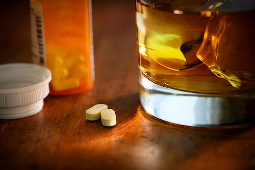 On a table is an open bottle of prescription pills with 2 on the table next to an alcoholic beverage in a glass.