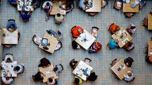 Bird's eye view of a dining hall with people sitting at square tables.